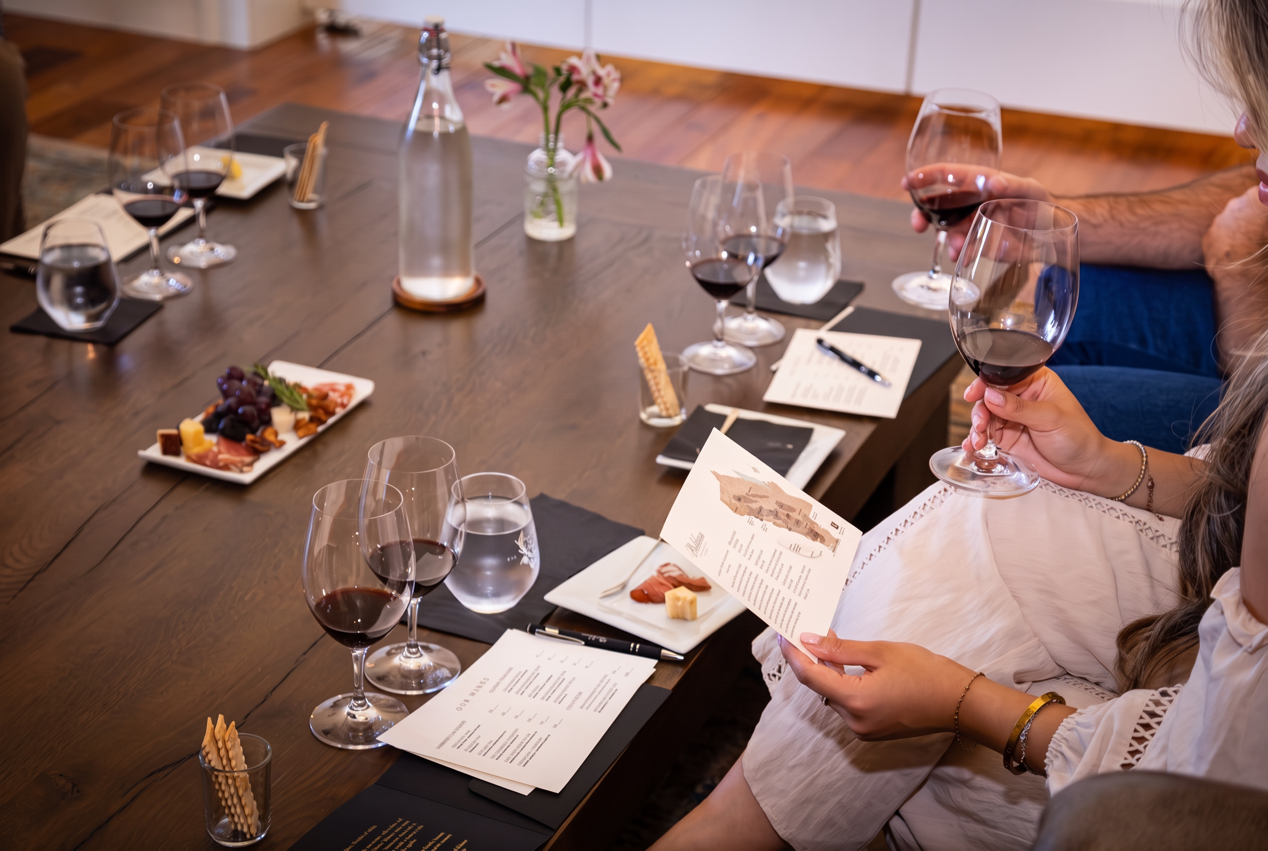Guests studying a tasting flight menu beside pours of red wine and a charcuterie plate